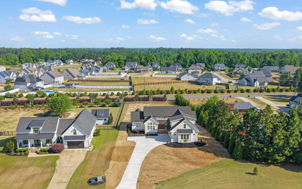 Aerial view of a suburban neighborhood with rows of houses, roads, and green lawns under a clear blue sky.