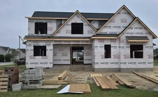 Partially built two-story house under construction with roofing, windows, and lumber in the yard