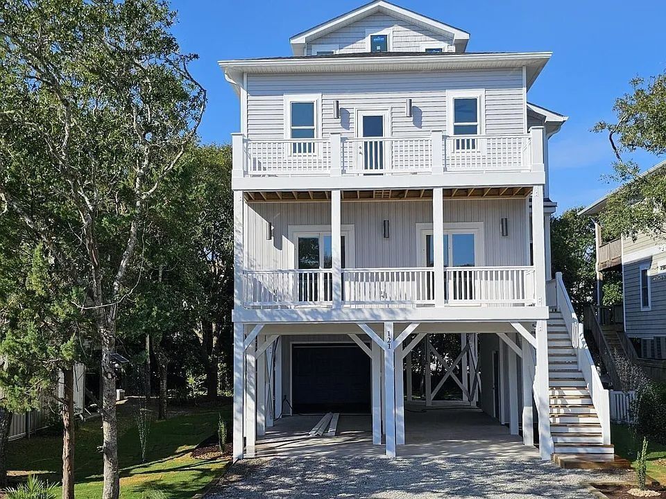 White two-story beach house with balconies, blue shutters, and stairs on both sides, surrounded by trees