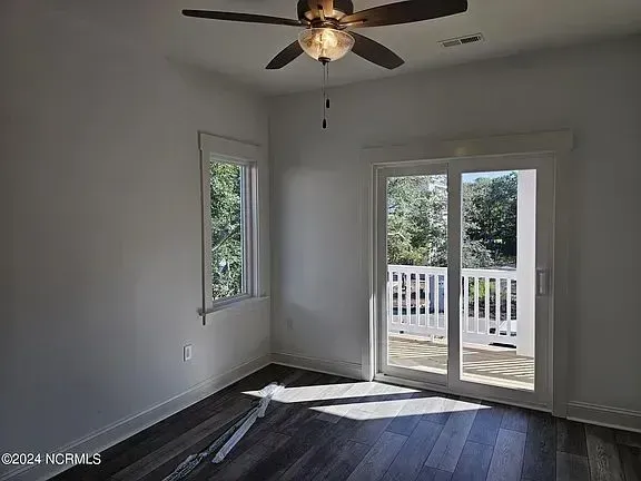 Empty room with dark wood floors, ceiling fan, window, and sliding glass door to a balcony.