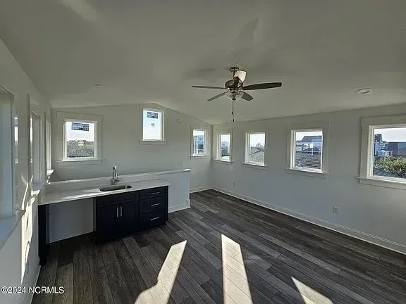 Bright empty kitchen with white walls, dark cabinets, and large windows over gray wood flooring.