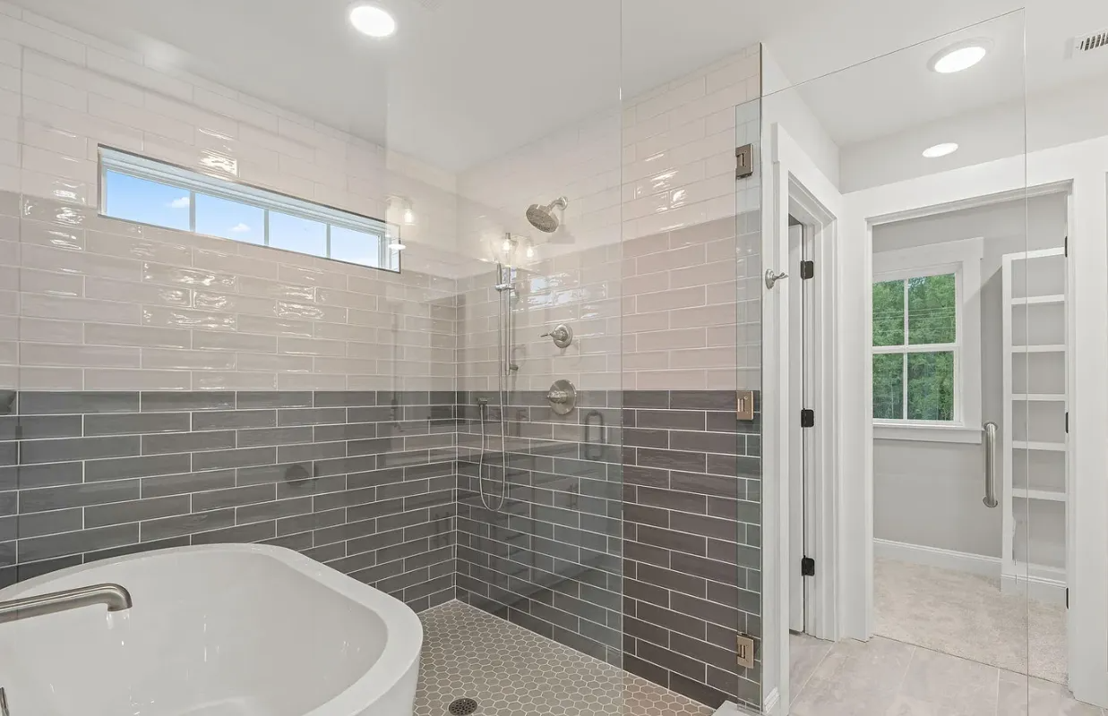 Modern bathroom with white tub, gray tile walls, and doorway to a bright adjoining room