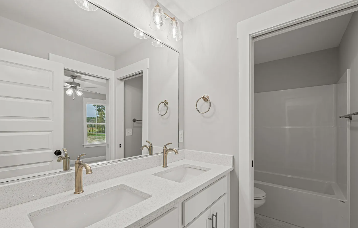 Bright white bathroom with double vanity, gold faucets, mirror, and a shower/tub alcove.