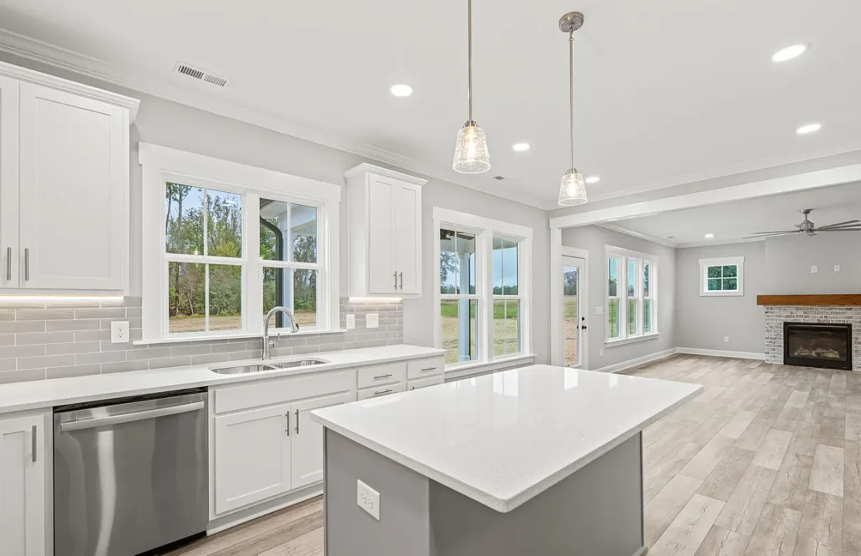 Bright modern white kitchen with island, stainless dishwasher, pendant lights, and open view to a fireplace living area
