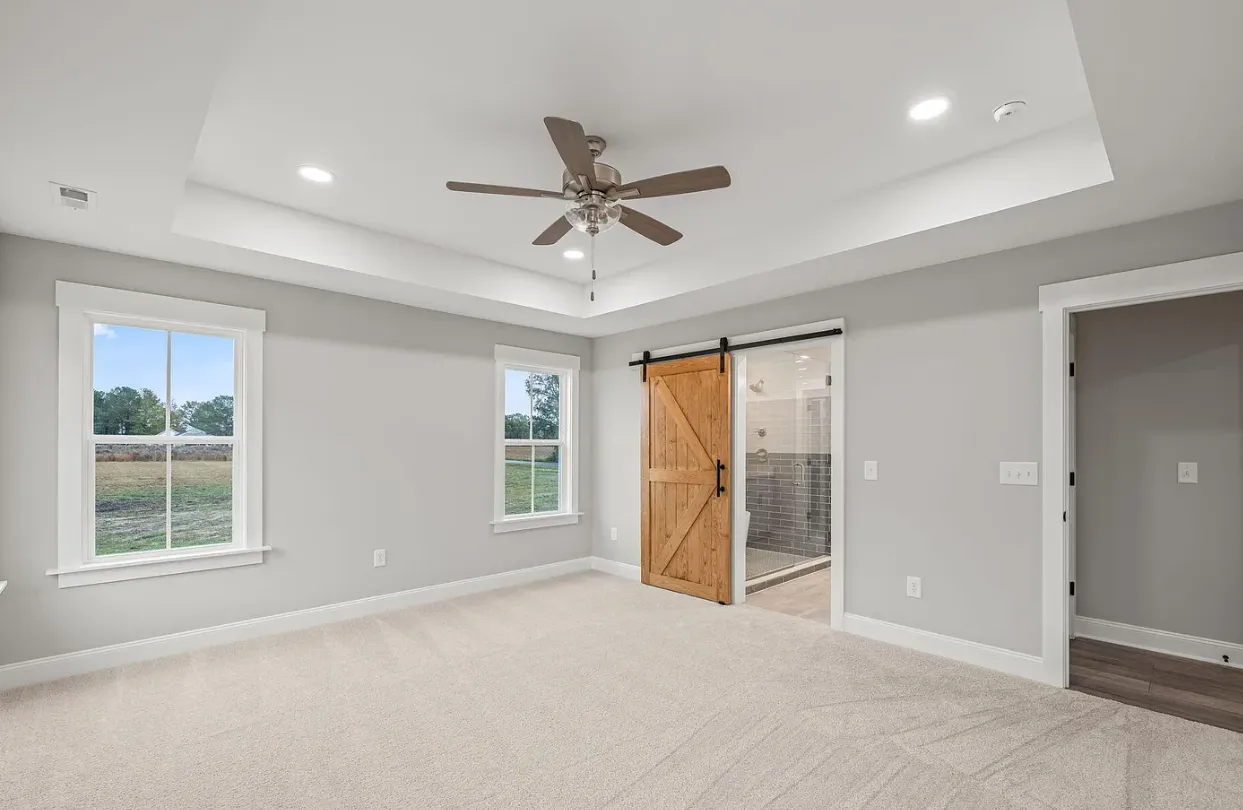 Spacious empty bedroom with beige carpet, gray walls, white trim, ceiling fan, and barn door to closet