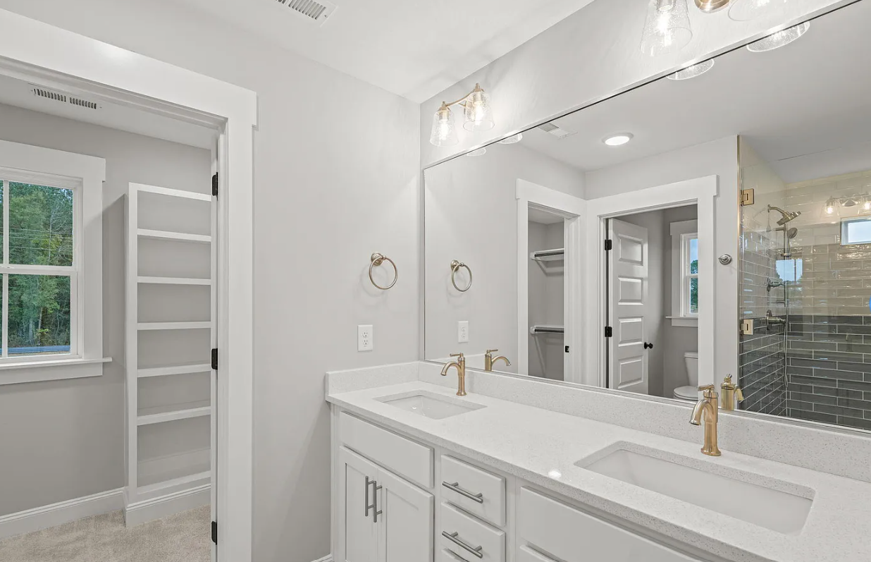 Bright white bathroom with double vanity, large mirror, and shower visible through an open doorway.