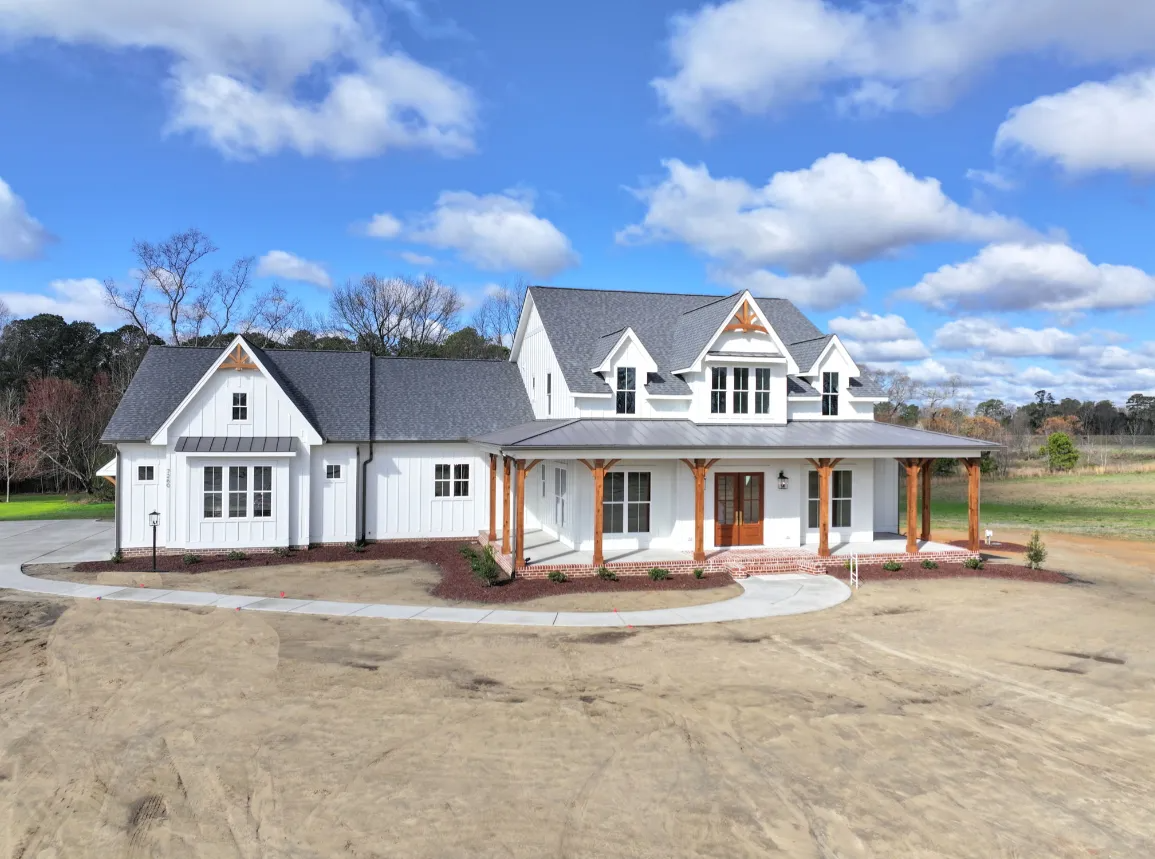 Large white house with a porch and gabled roof on a dirt lot under a partly cloudy sky