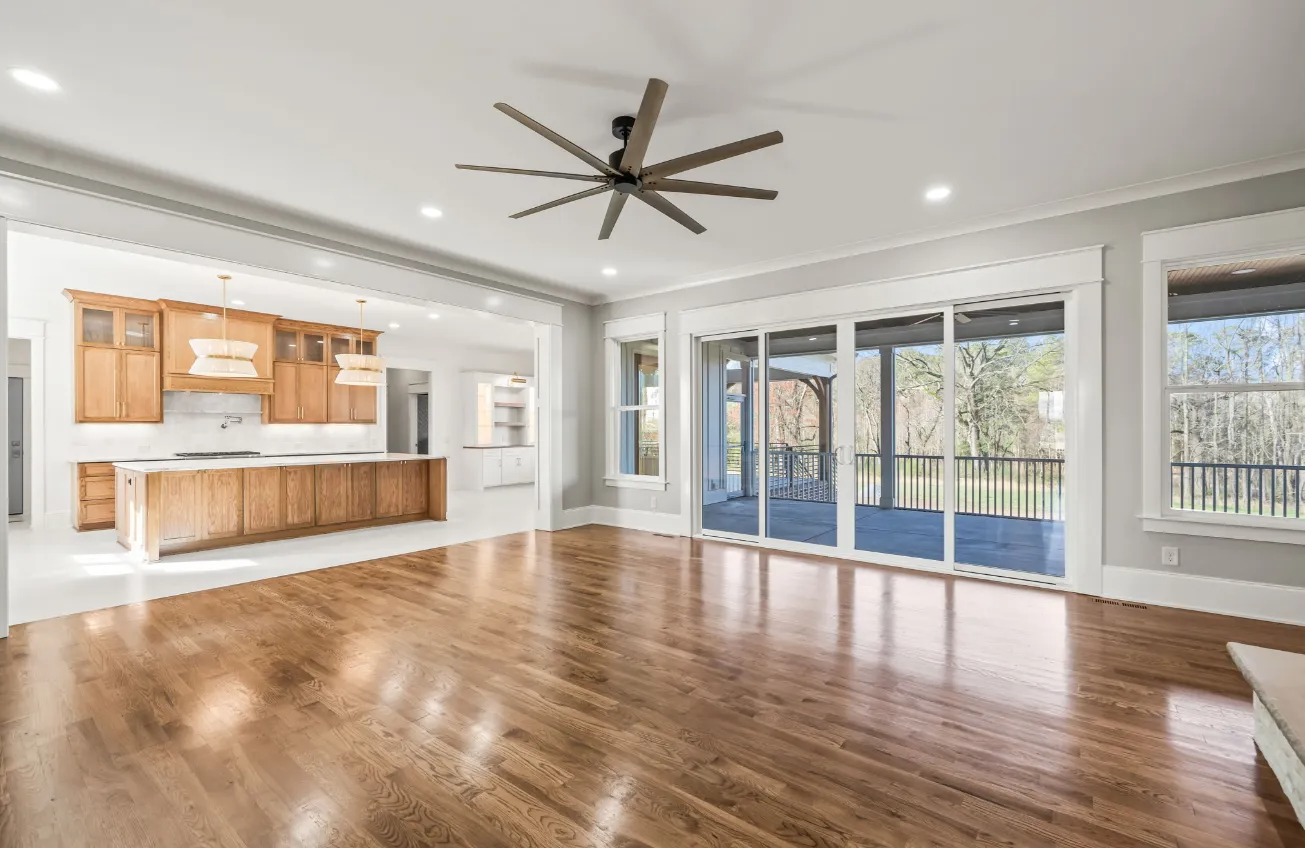 Spacious empty living room with hardwood floors, ceiling fan, and open view to kitchen and large windows.