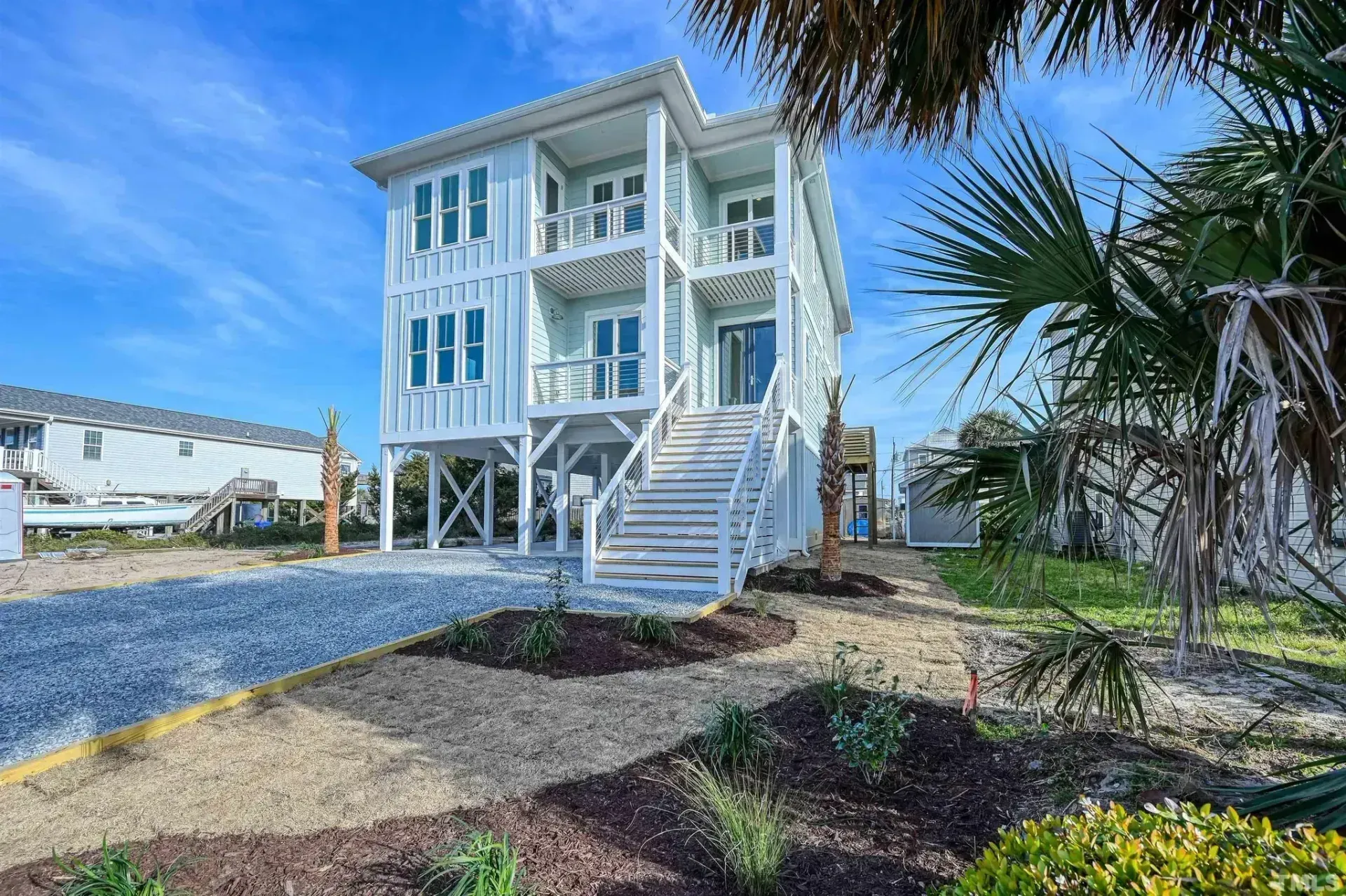 White elevated beach house with wraparound porches and front stairs under a blue sky, surrounded by landscaping