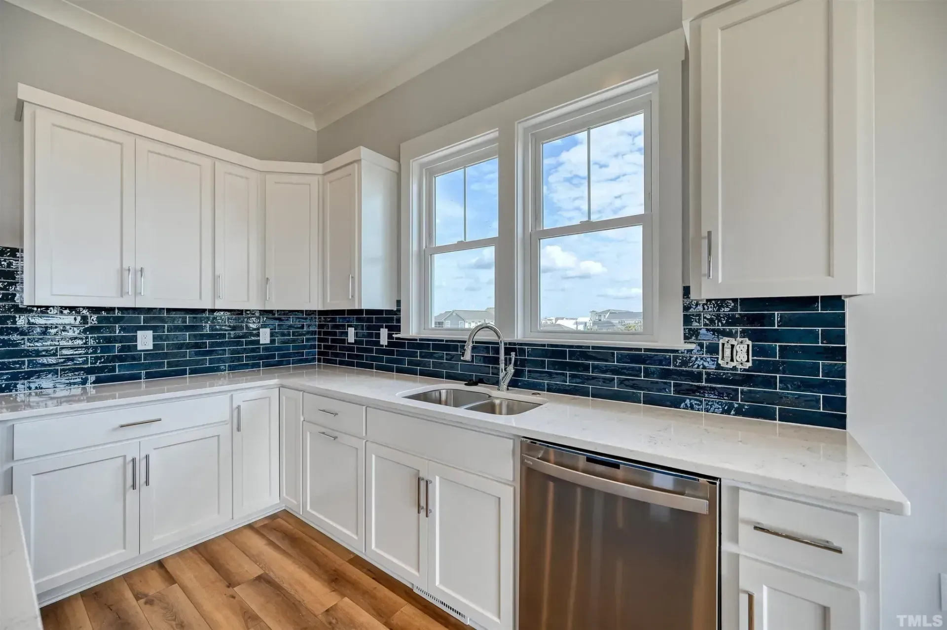 Bright white kitchen with blue tile backsplash, wood floor, and stainless steel dishwasher under a window.