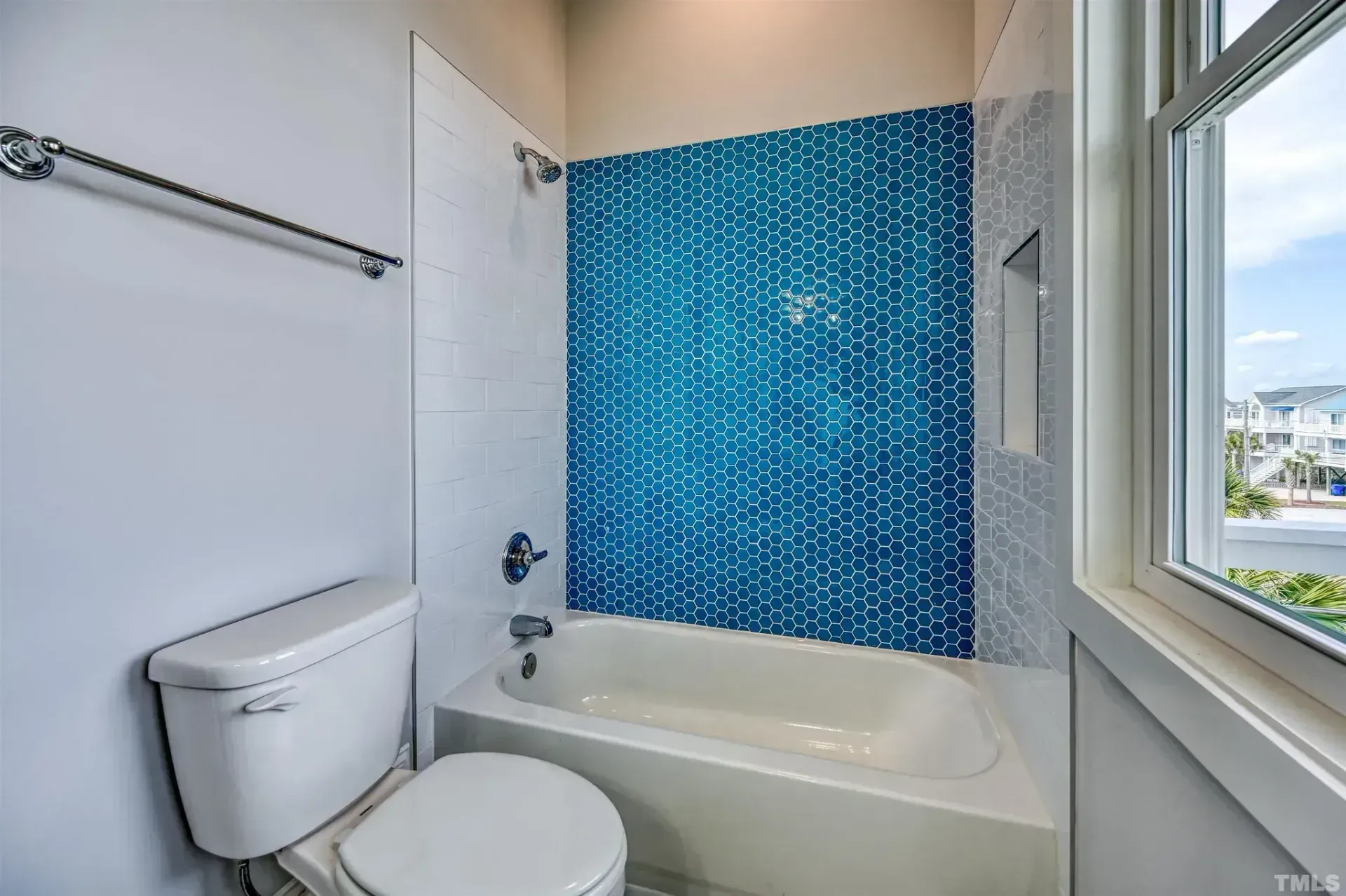 Small bathroom with white tub, toilet, and blue mosaic tile shower wall beside a window.
