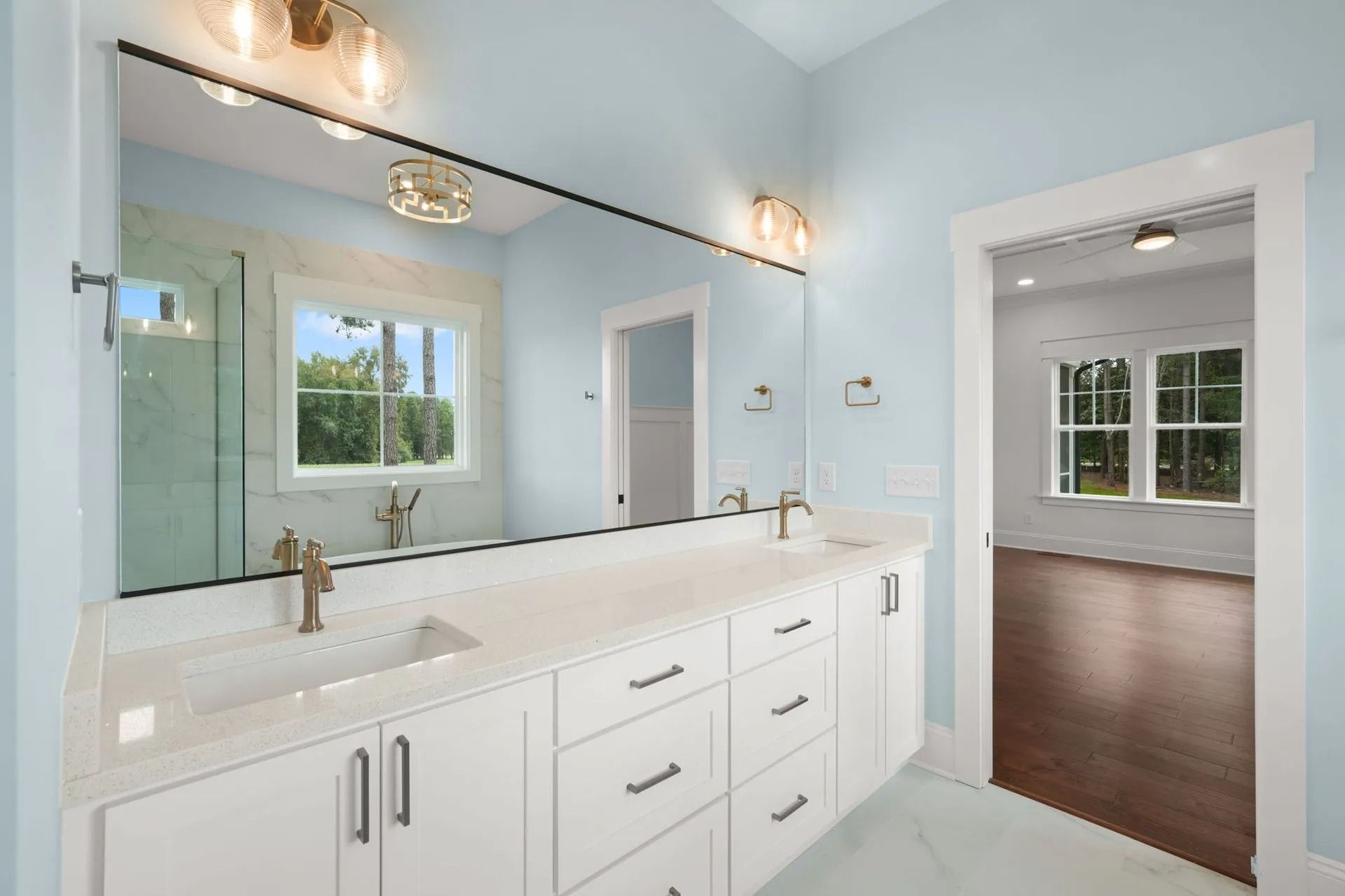 Bright modern bathroom with white vanity, large mirror, and a doorway to a bedroom with hardwood floors