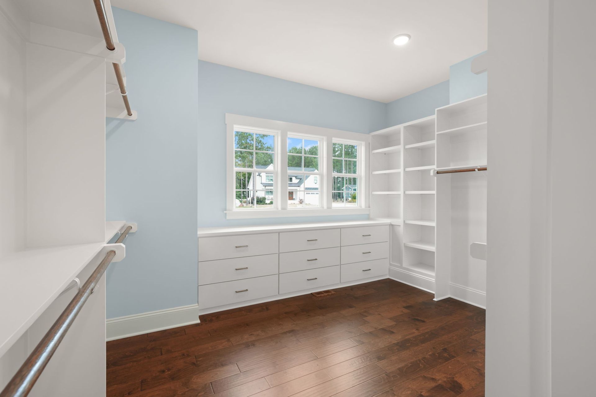 Bright empty walk-in closet with white shelves, drawers, and windows over dark wood floors.