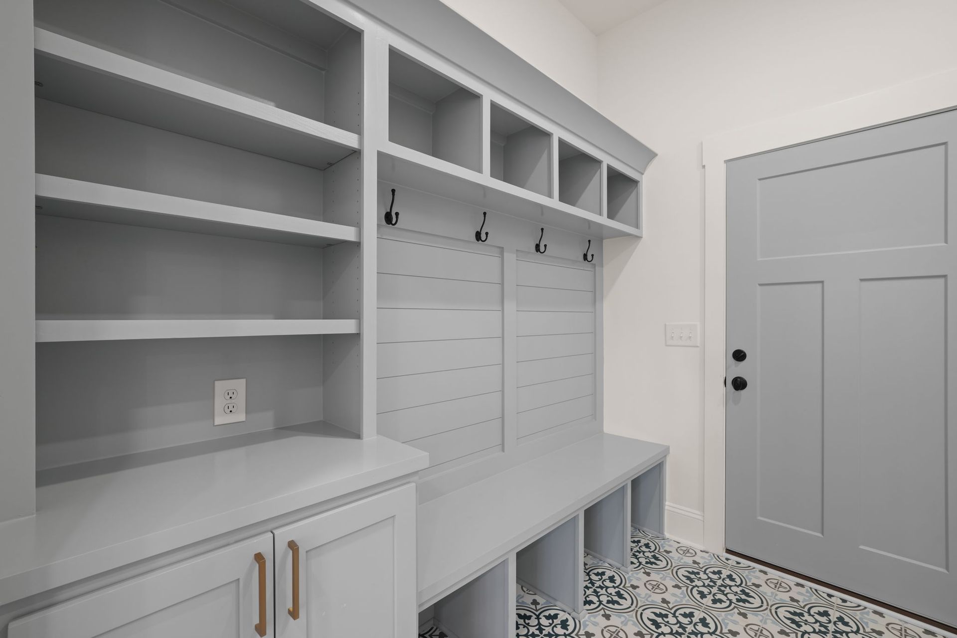 Bright mudroom with white built-in cabinets, hooks, drawers, and patterned tile floor by a gray door