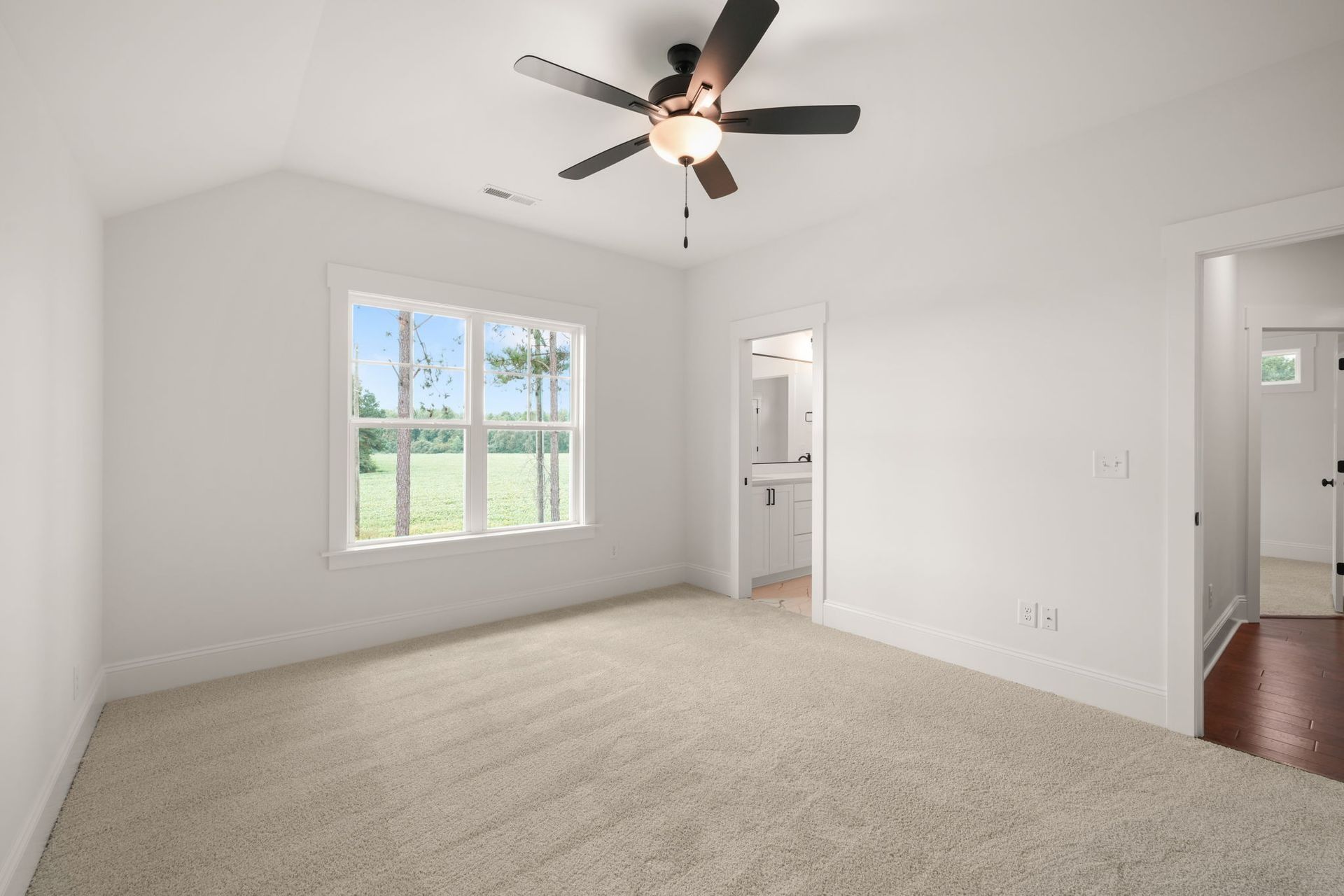 Empty carpeted bedroom with ceiling fan, white walls, and a window overlooking greenery