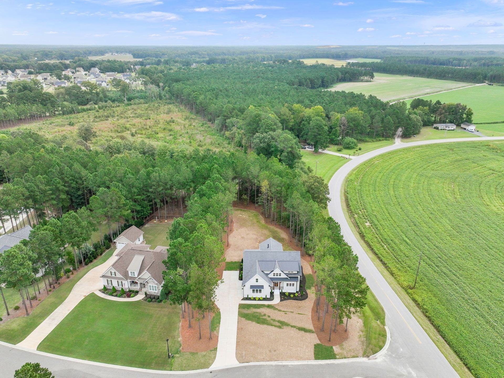 Aerial view of two houses beside a curved road, surrounded by green fields and trees.