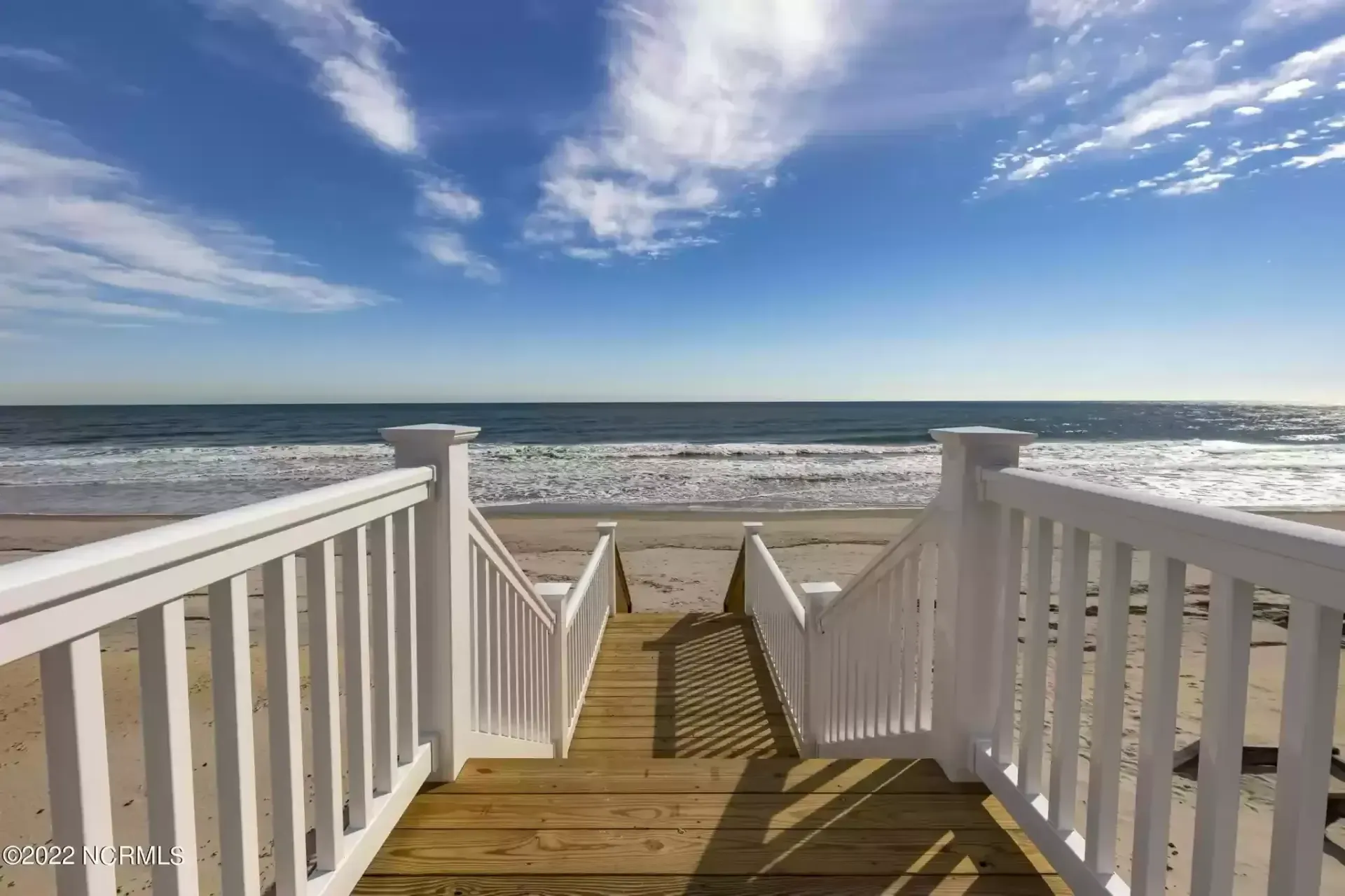 White wooden stairs leading to a sandy beach and ocean under a bright blue sky