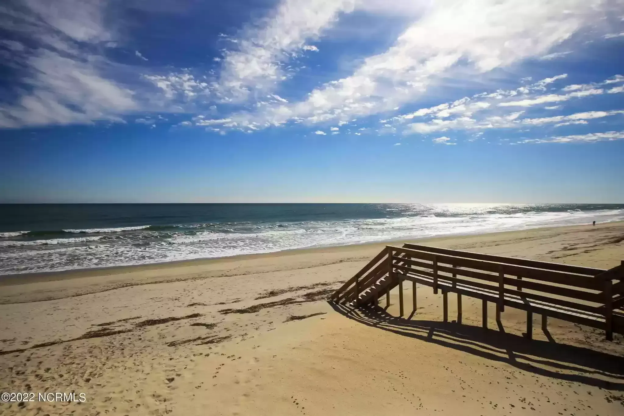 Sunny beach with blue sky, ocean waves, and a wooden stairway descending onto the sand