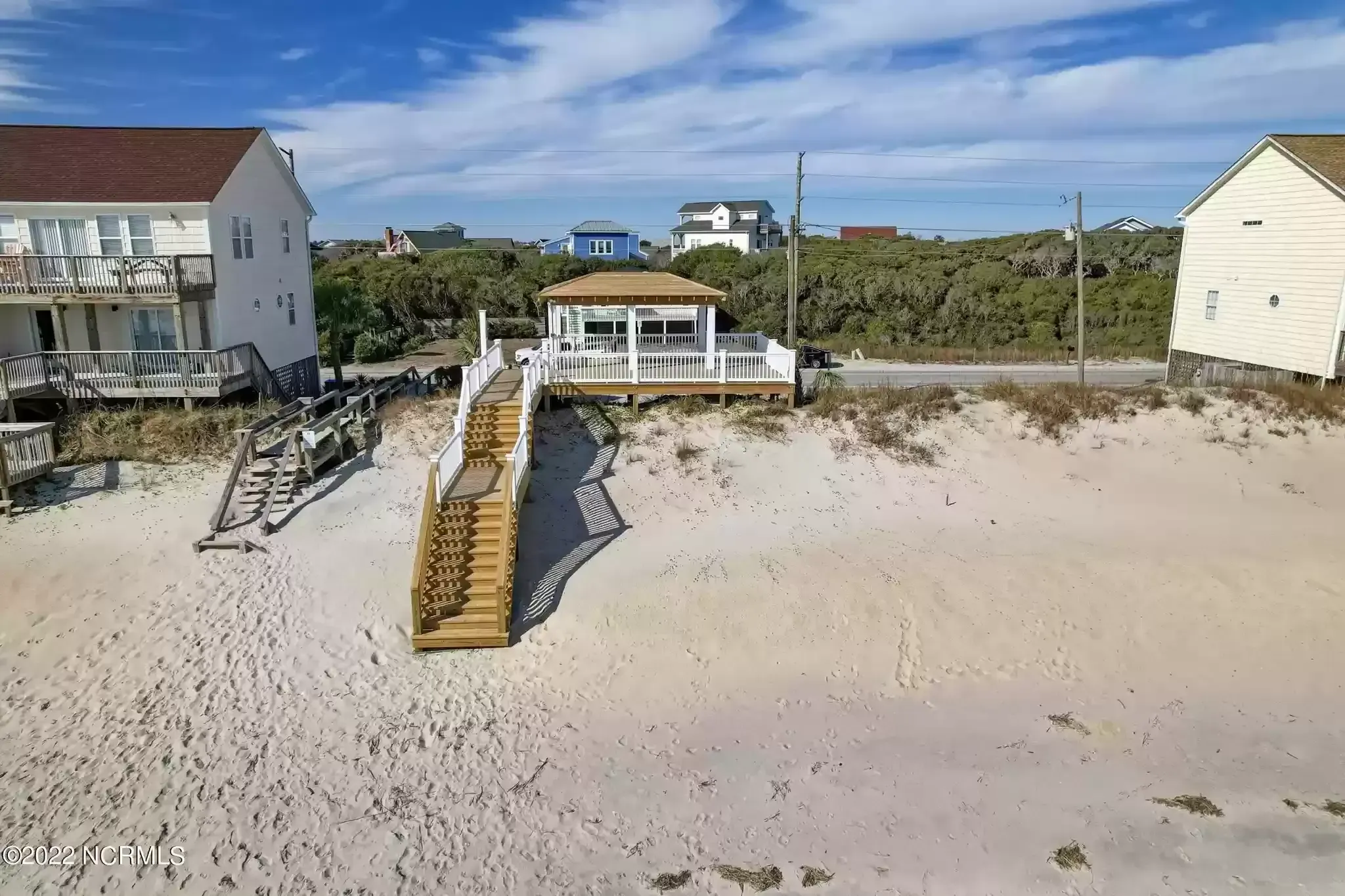 Beachfront houses above a sandy shore with a wooden stairway and blue sky