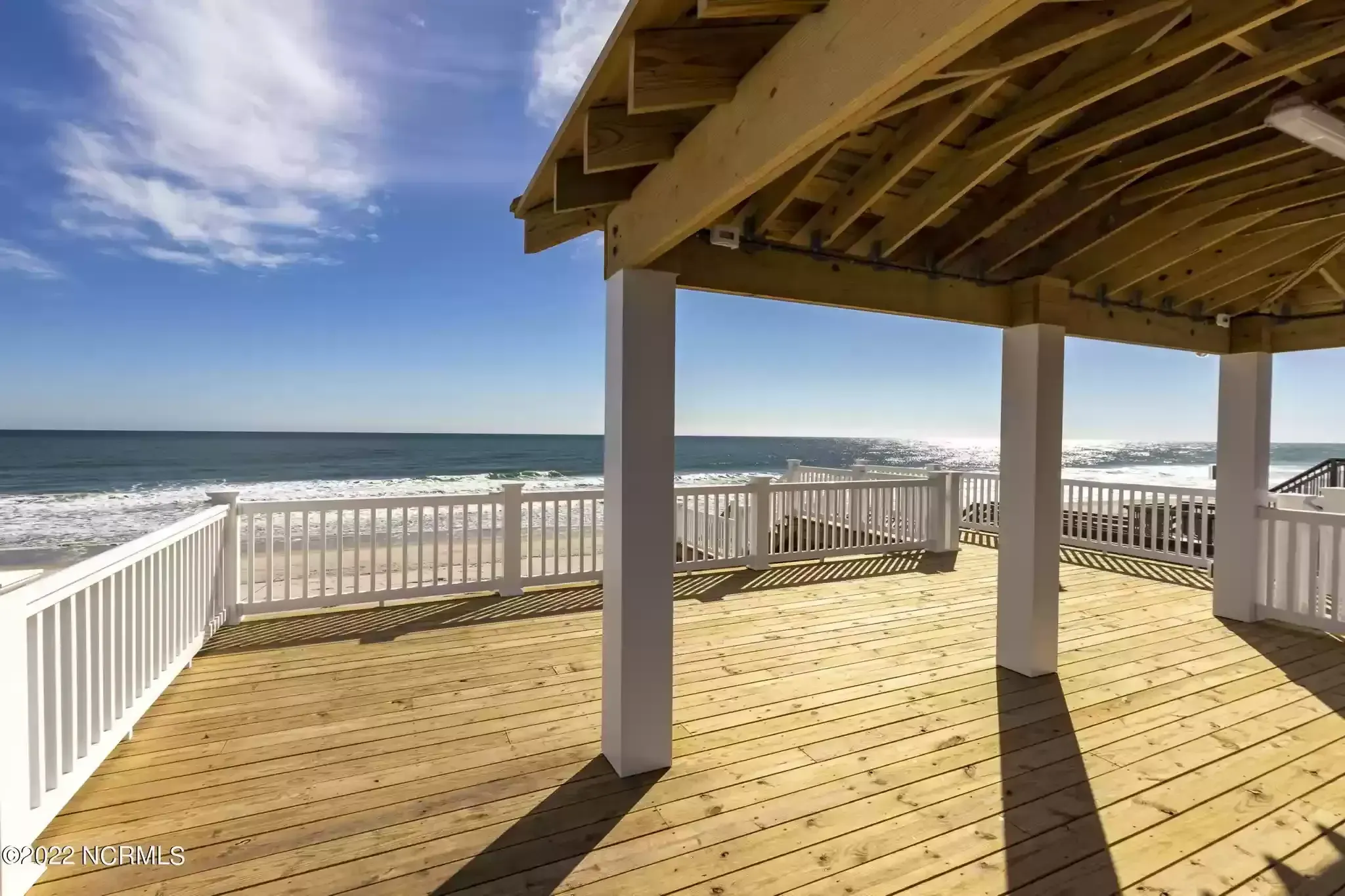 Ocean-view deck with white railing, wooden floor, and covered pergola under a blue sky