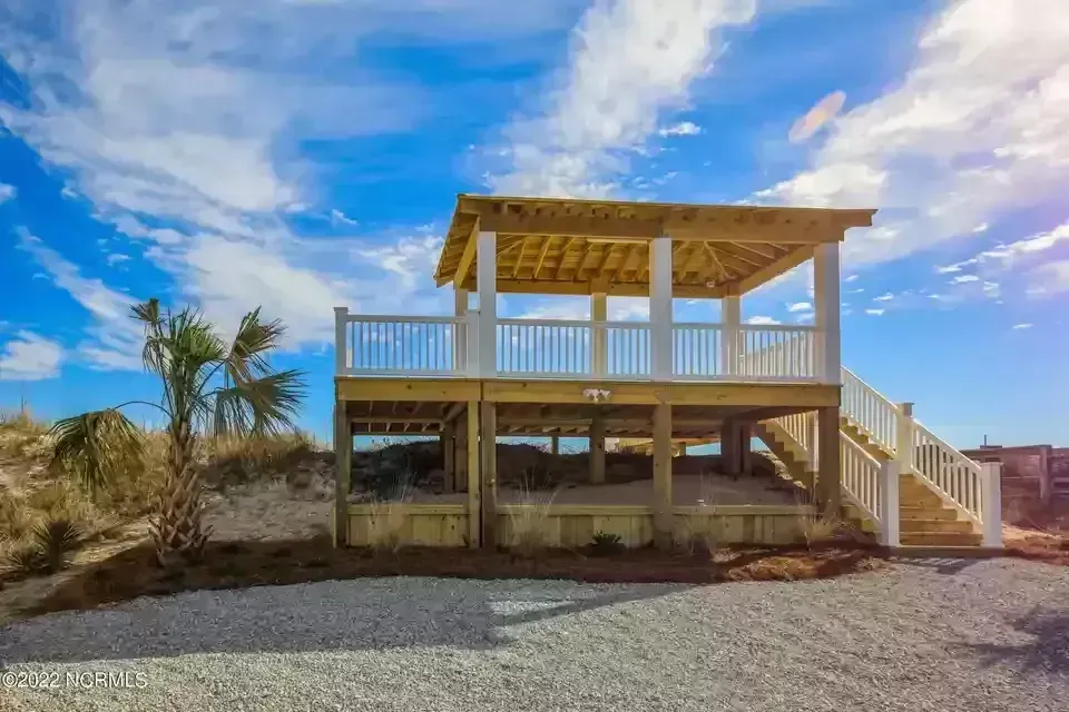 Two-story beach pavilion with a rooftop deck, stairs, and palm trees under a blue sky
