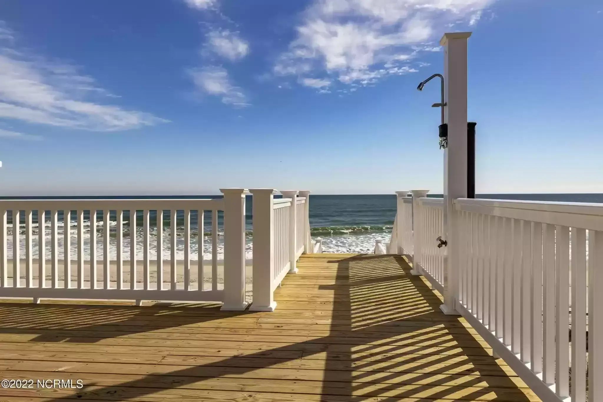 Sunny beach boardwalk with white railings leading to the ocean under a blue sky.