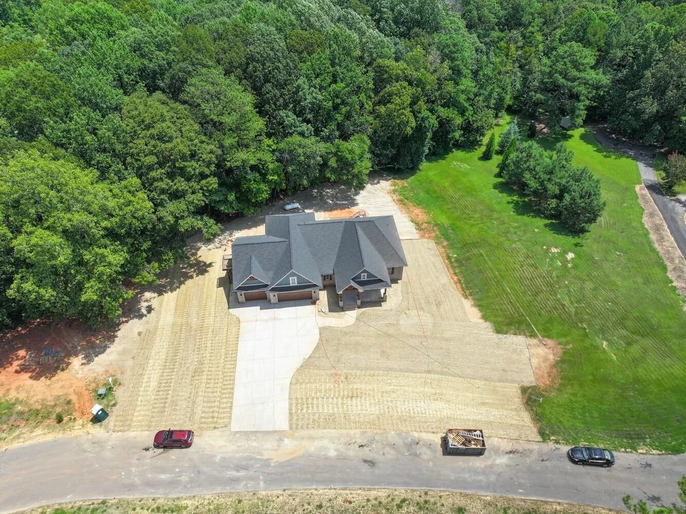 Aerial view of a house under construction with dirt lot, green trees, and parked vehicles nearby
