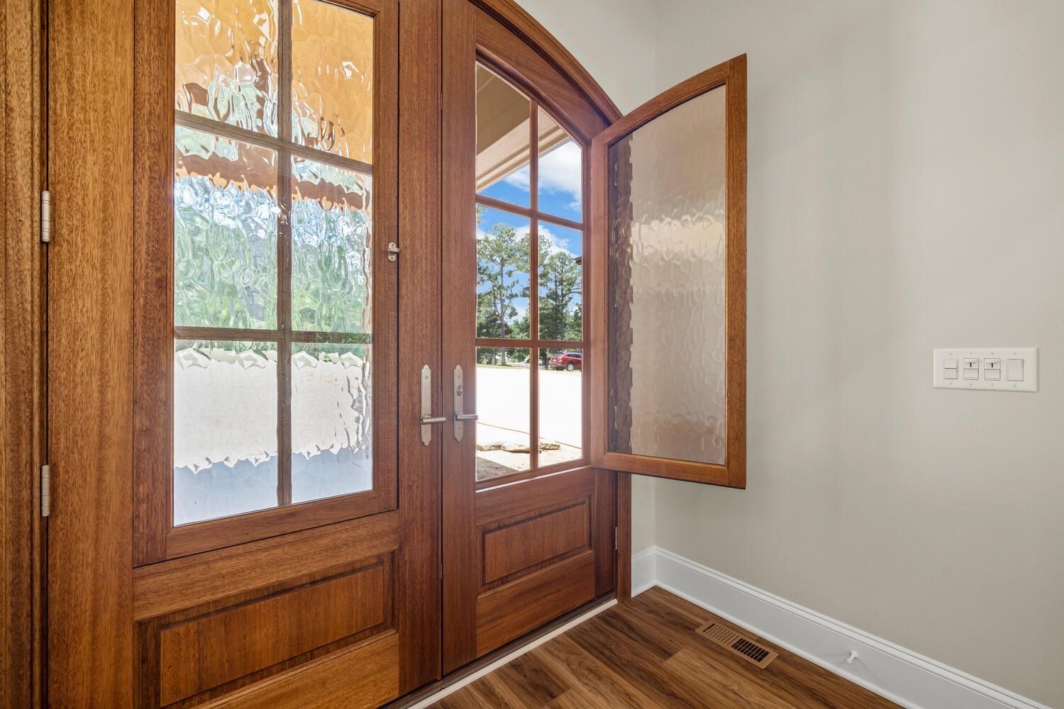 Wooden front door with frosted glass sidelights, open window, and hardwood floor in a bright entryway