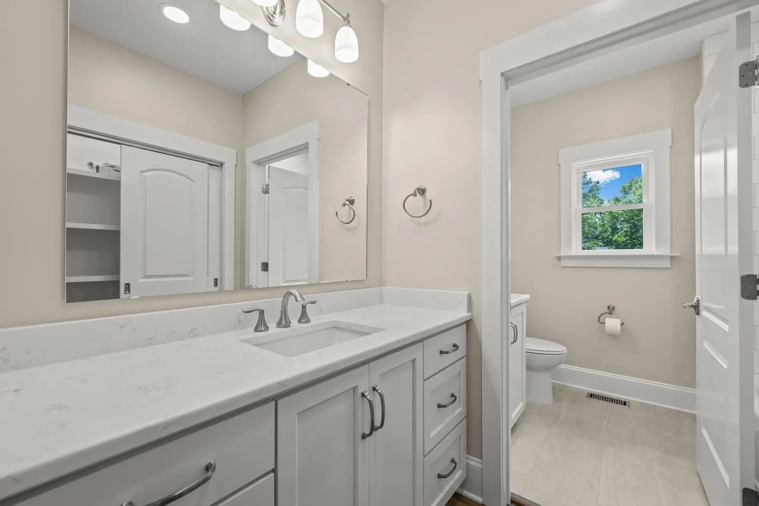 Bright bathroom with double vanity, large mirror, and toilet beside a window.