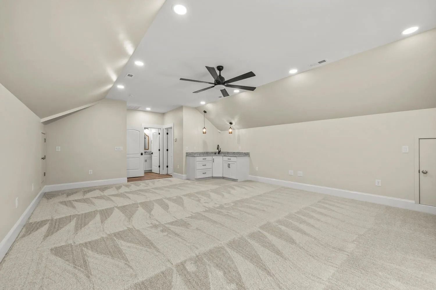 Spacious empty room with vaulted ceiling, ceiling fan, light carpet, and a small kitchenette area.