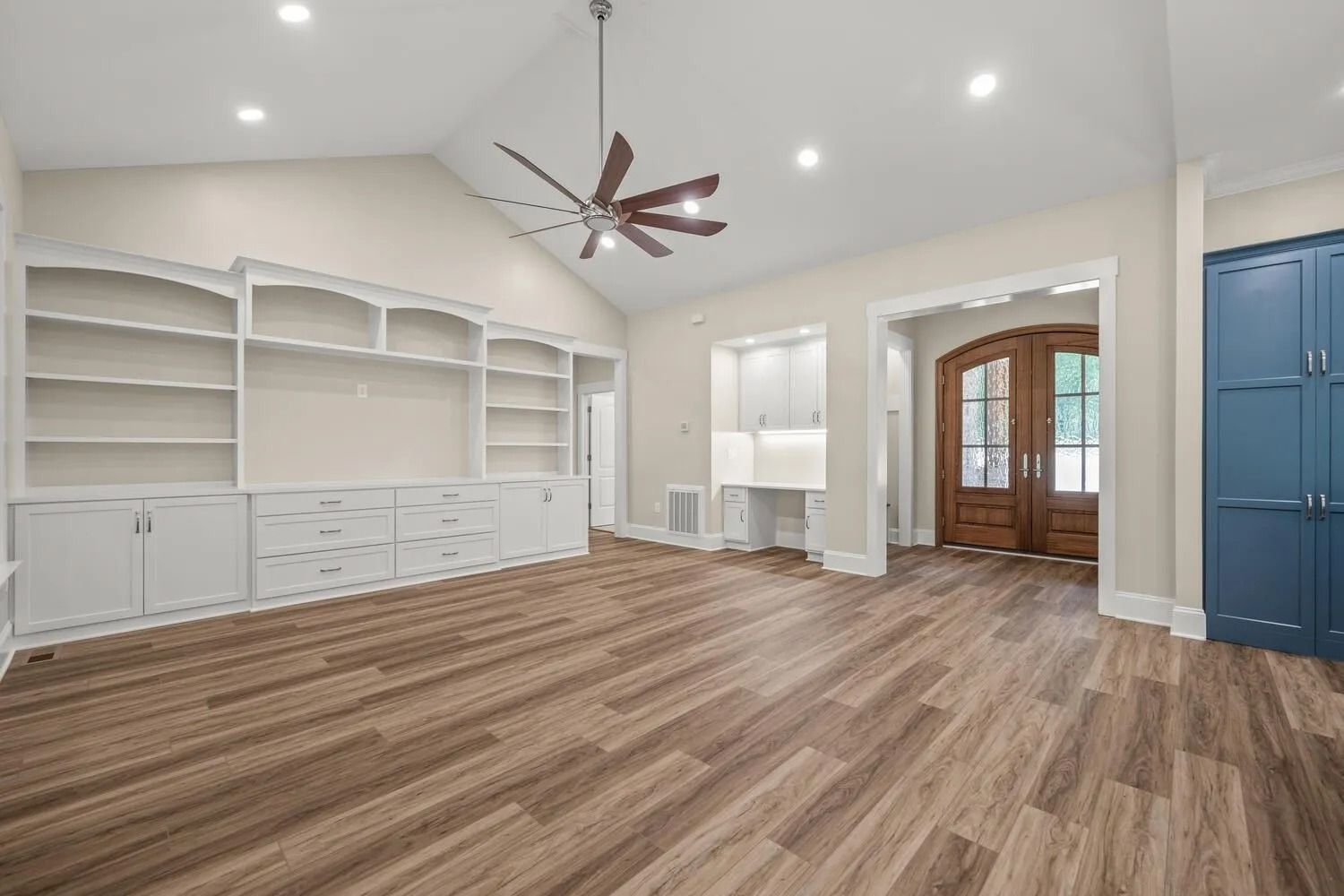 Bright empty room with white built-in shelving, wood floors, ceiling fan, and French doors
