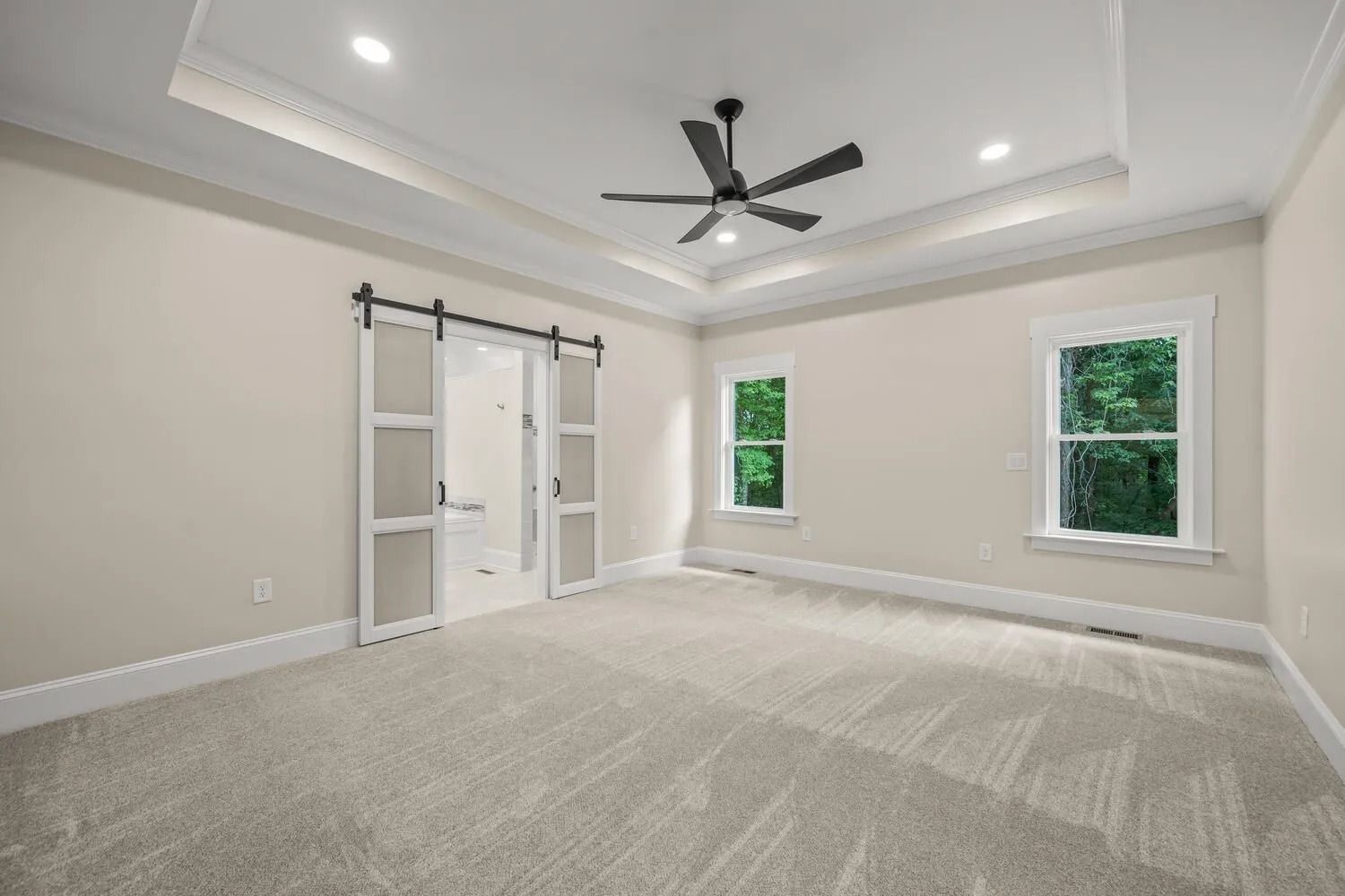 Empty modern bedroom with gray carpet, white walls, ceiling fan, barn doors, and two windows.