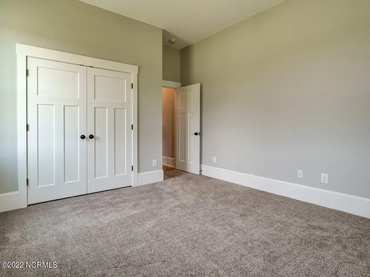 Empty beige bedroom with carpet, white double closet doors, and an open door to a hallway.
