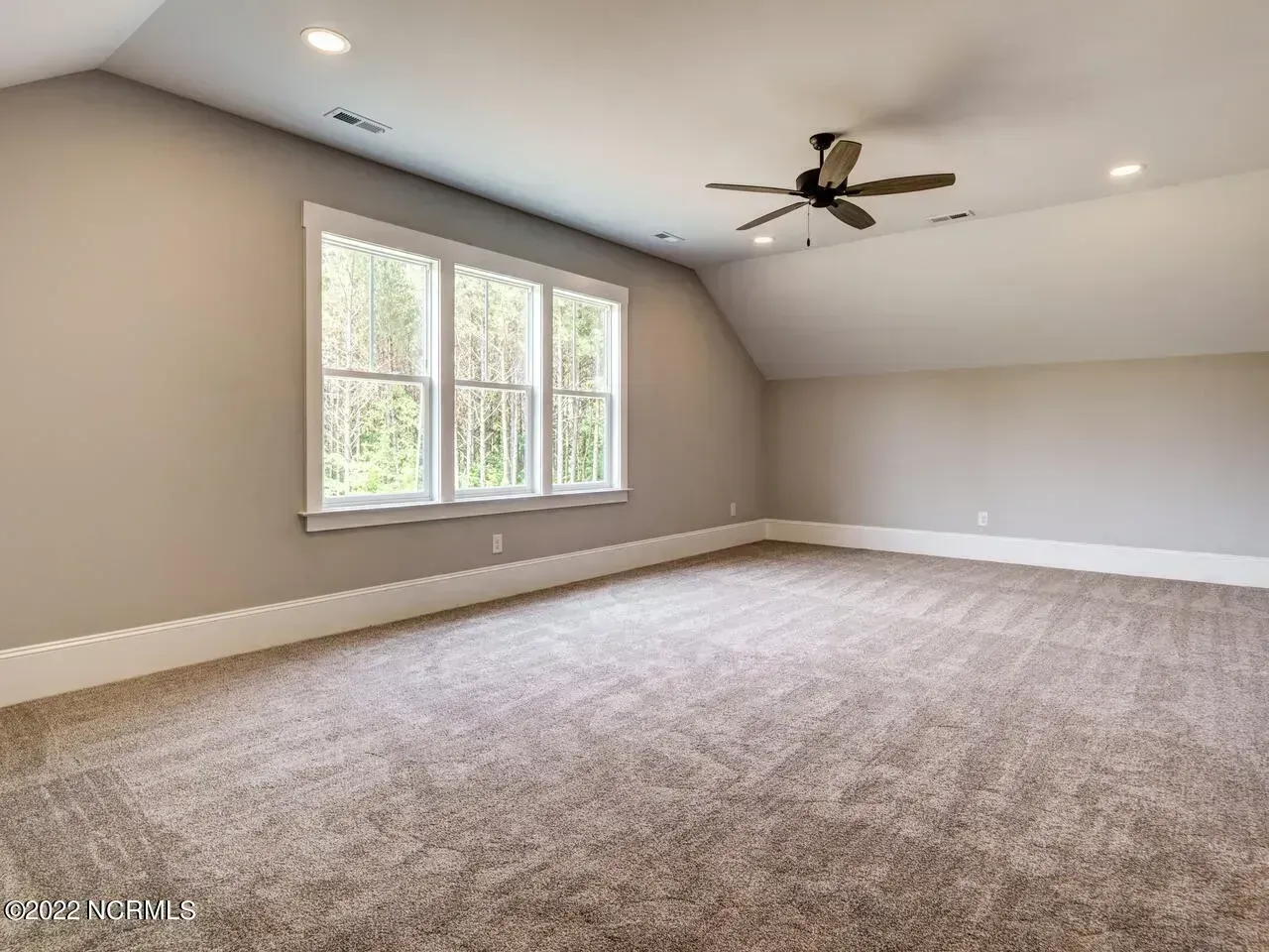 Empty carpeted room with sloped ceiling, window, and ceiling fan in neutral tones