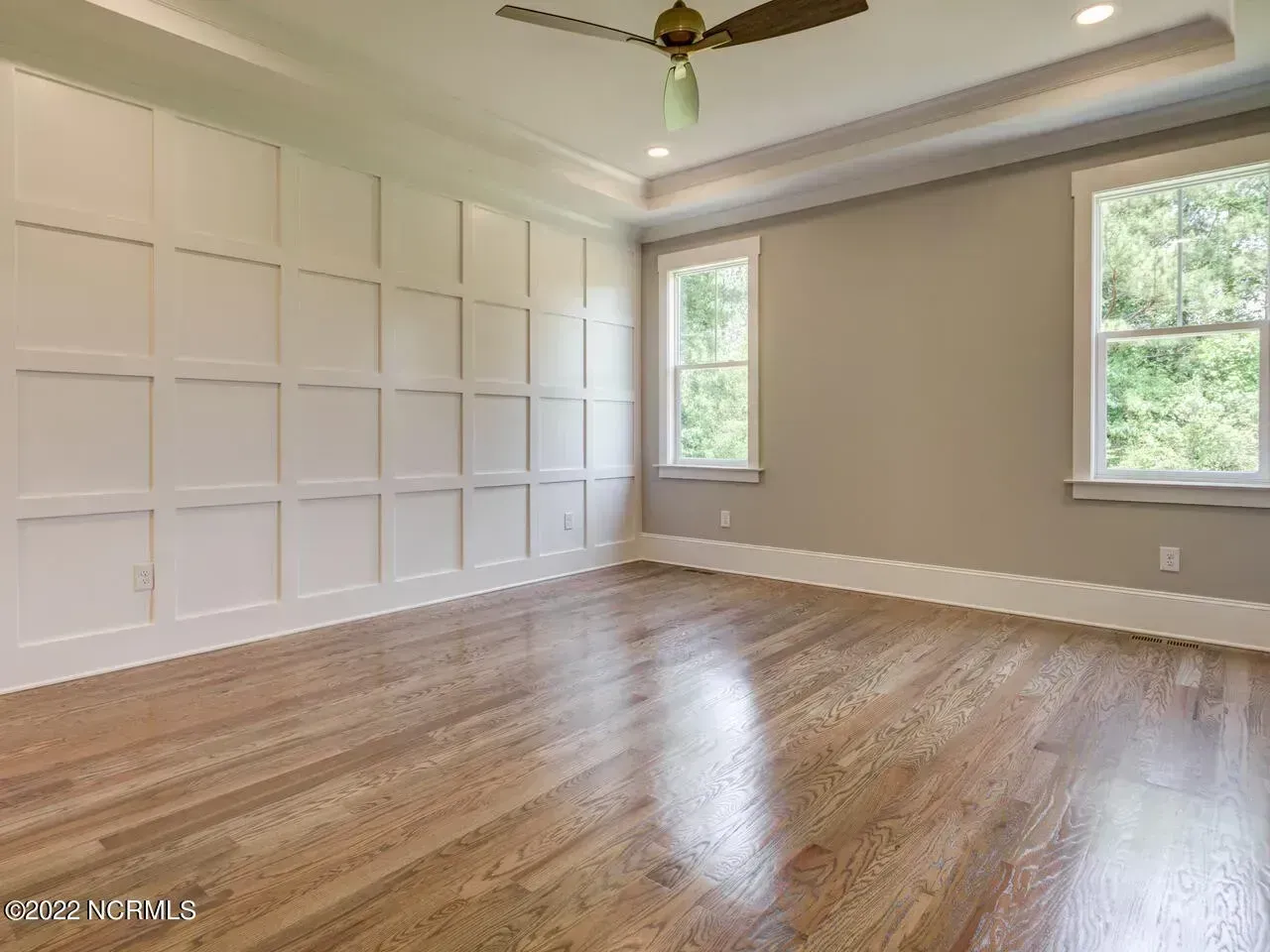 Empty living room with white paneled wall, hardwood floors, ceiling fan, and two windows