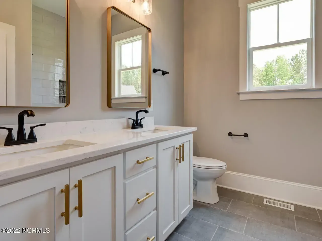 Bright modern bathroom with white vanity, black fixtures, gold handles, and a toilet by a window.