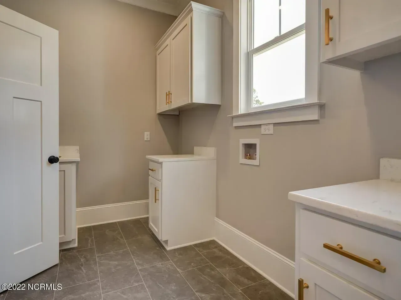 Small laundry room with white cabinets, gray tile floor, and a window letting in daylight.