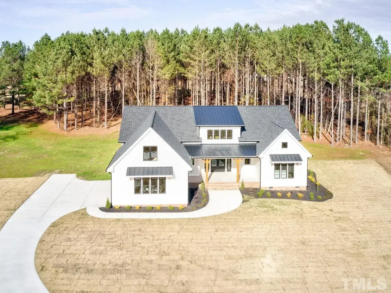 White two-story house with gray roof and large driveway, set in front of a pine forest