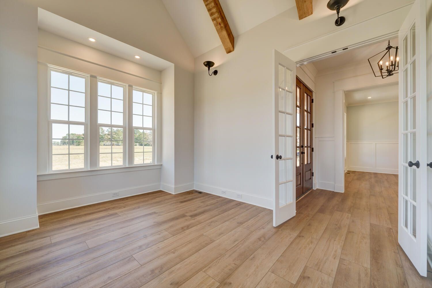 Bright empty room with wood floors, exposed beams, large windows, and open French doors