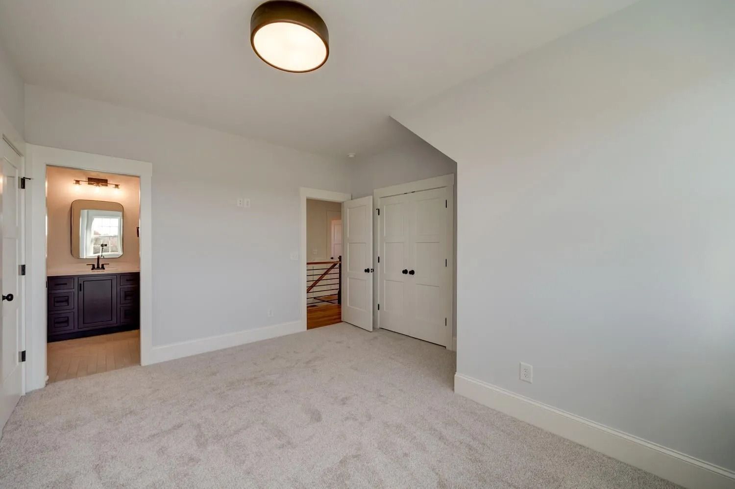 Empty carpeted bedroom with white walls, ceiling light, open bathroom door, and closet doors.