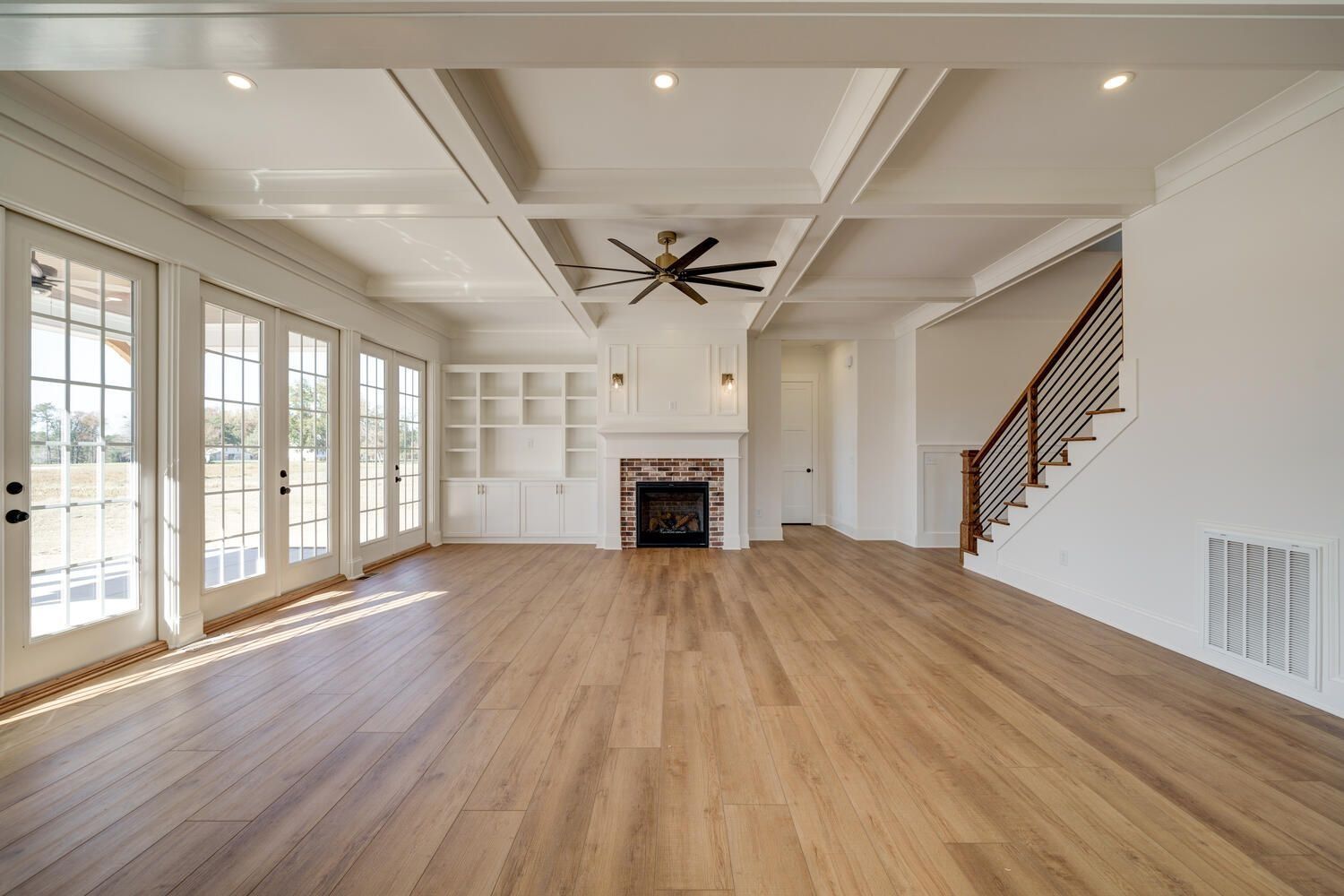 Bright empty living room with hardwood floors, fireplace, built-ins, large windows, and staircase