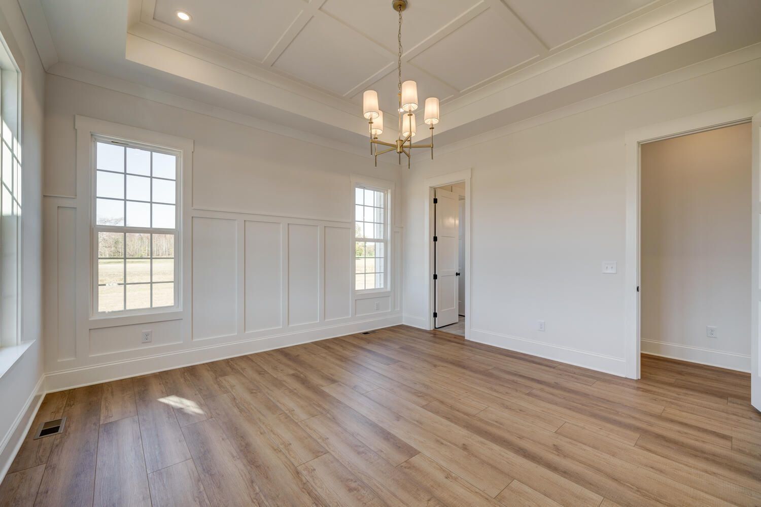 Empty bright room with hardwood floors, white walls, tall windows, and a chandelier-lit tray ceiling