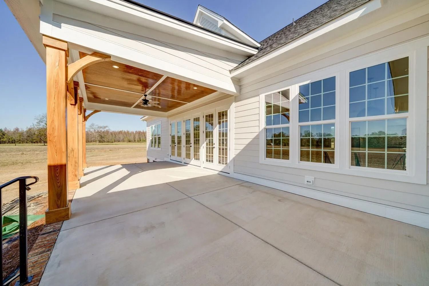 Covered concrete patio with white siding, large windows, and exposed wooden beams under a bright blue sky