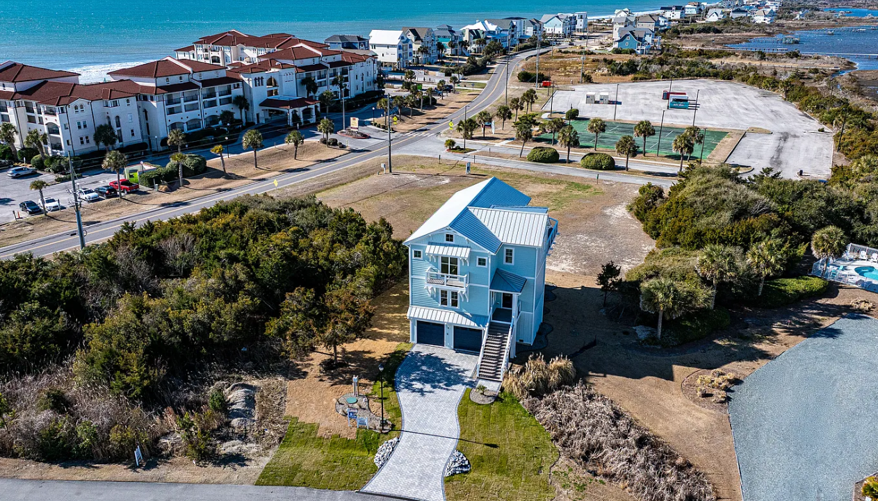 Blue waterfront house with a white walkway, surrounded by trees and dry land near a shoreline.