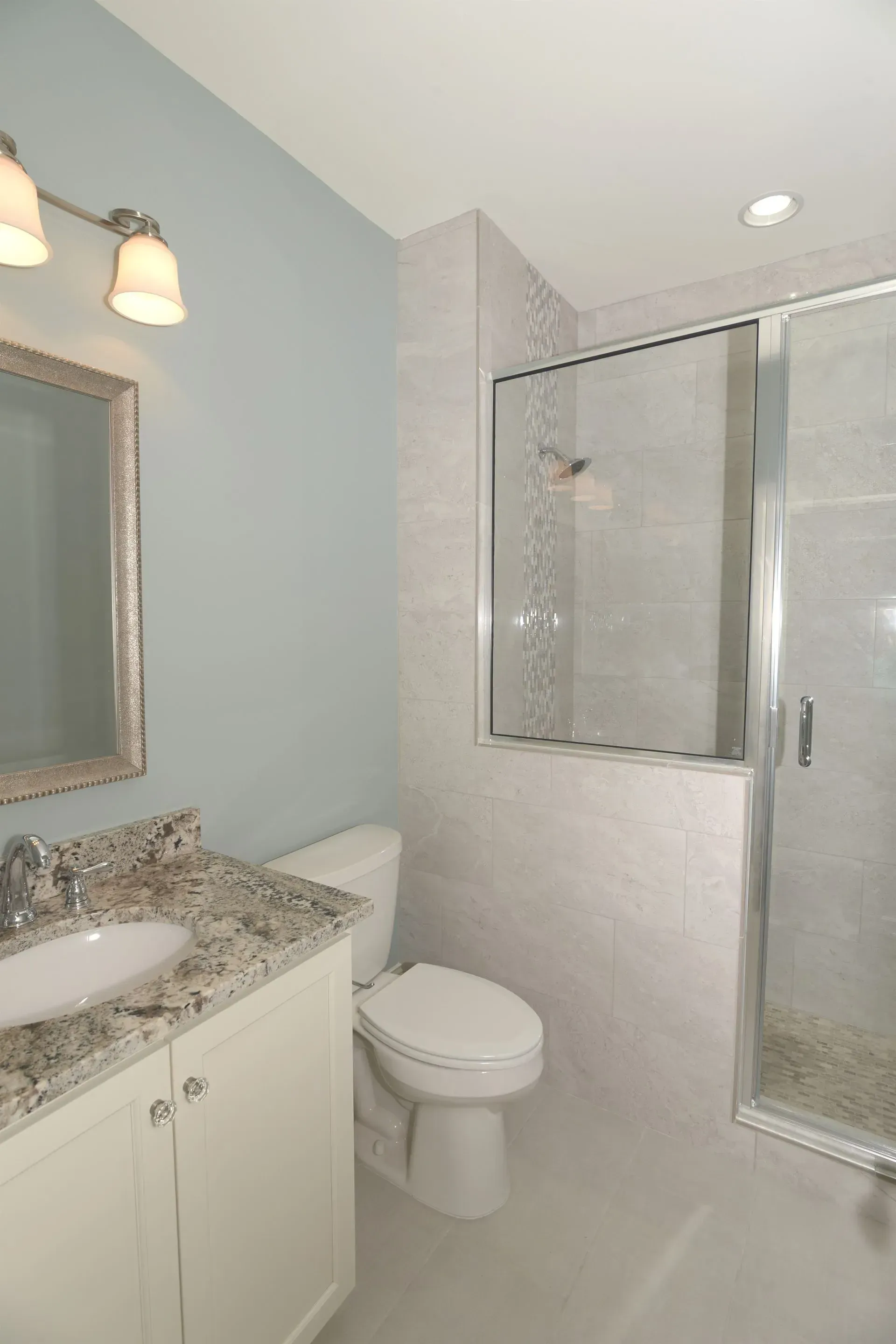 A bathroom featuring a light blue wall, a vanity with a granite countertop, a toilet, and a glass-enclosed shower.