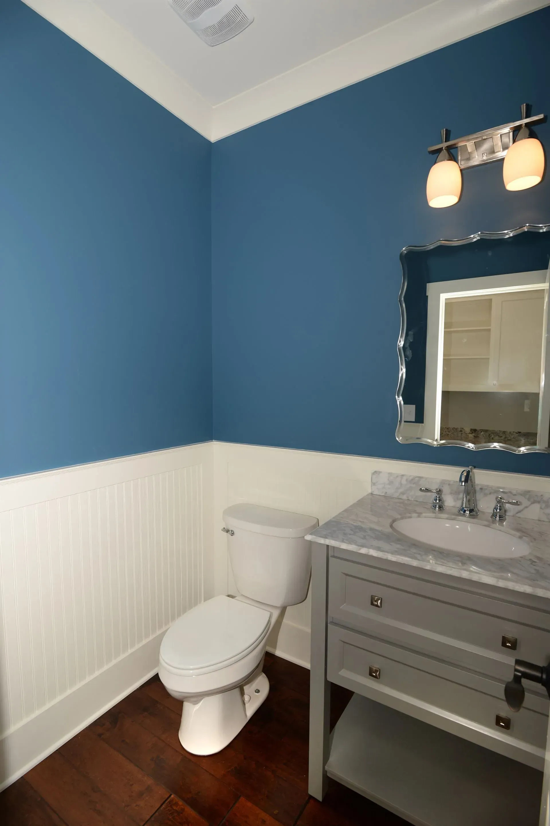 A half-bathroom with blue walls, white beadboard wainscoting, a pedestal vanity, and a toilet on dark wood flooring.