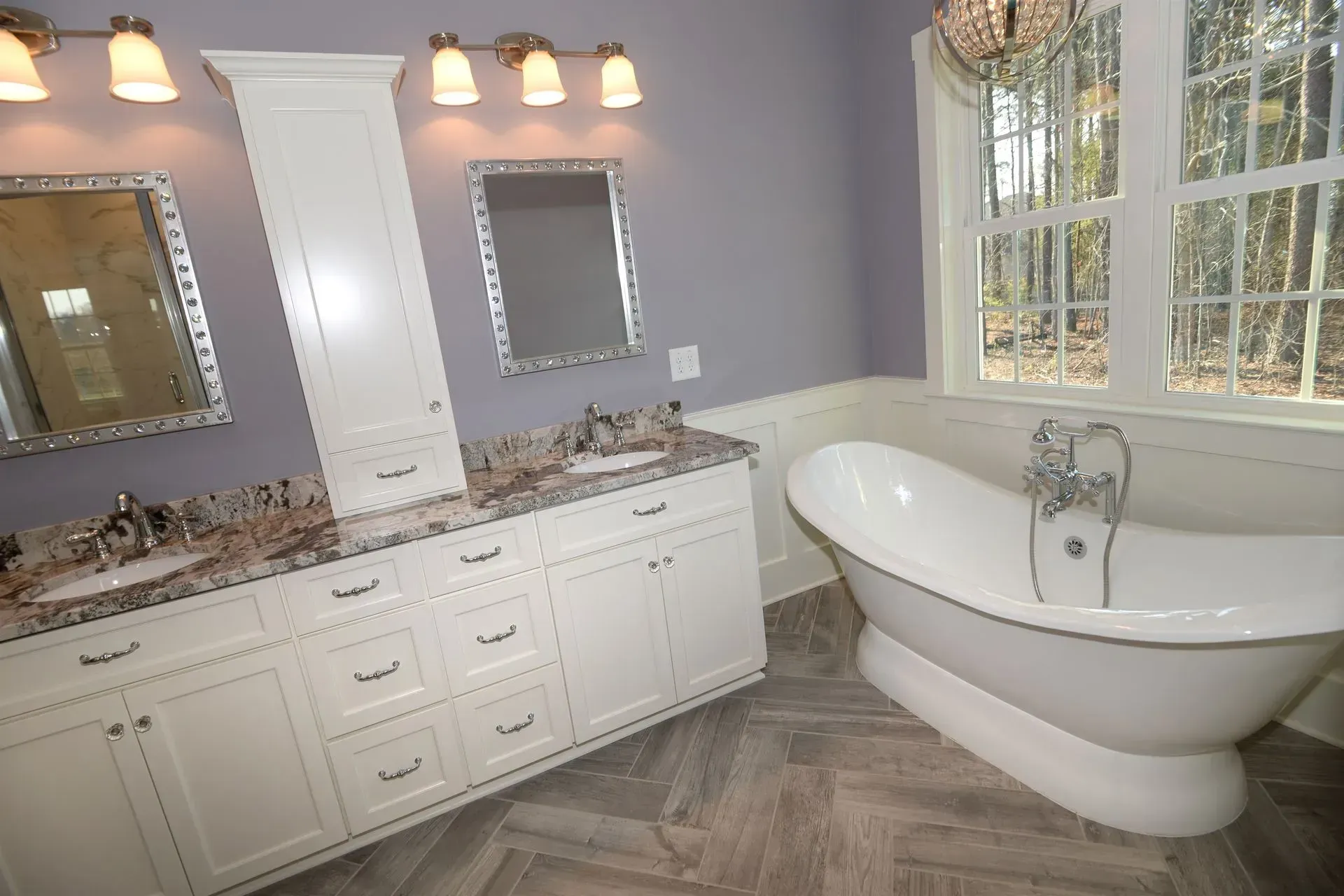 Modern bathroom with double vanity, white cabinetry, purple walls, herringbone floor, and a white freestanding bathtub.