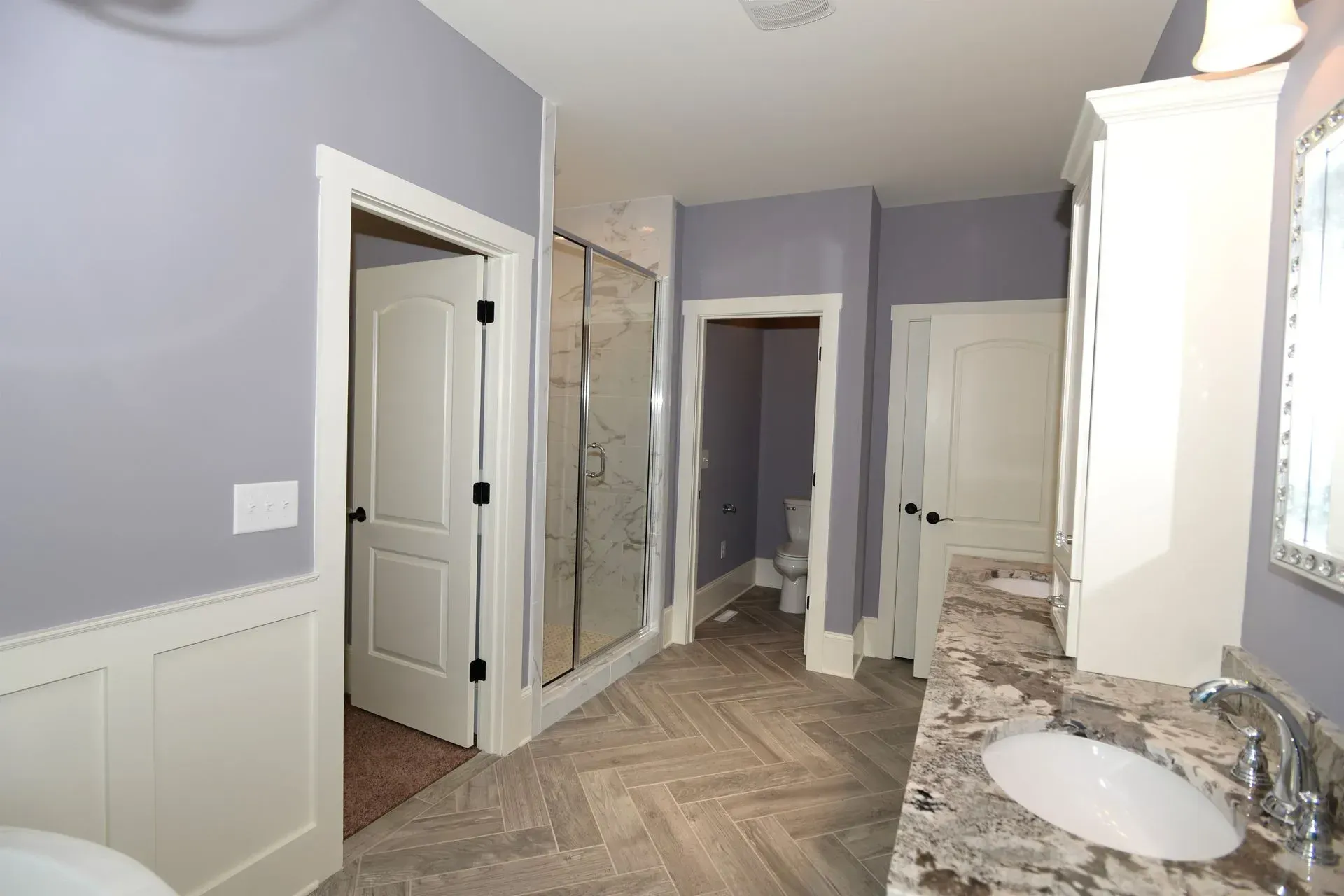 A bathroom featuring light purple walls, a granite countertop vanity, a glass-enclosed shower, and a herringbone floor.