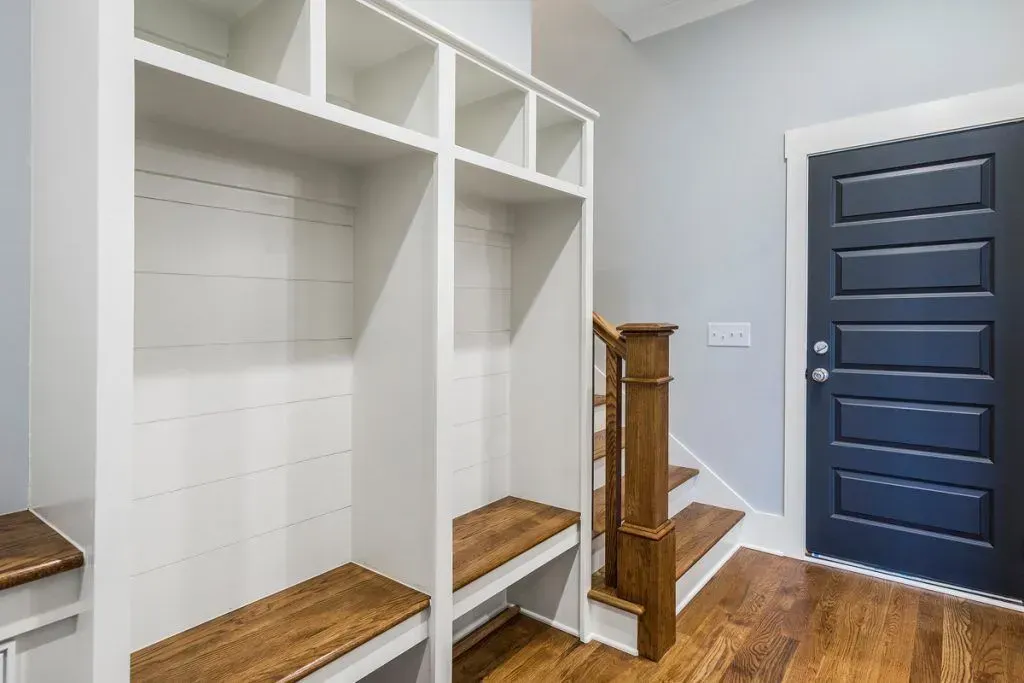 A white mudroom built-in unit with wood benches next to a dark blue door and a staircase with a wood railing.