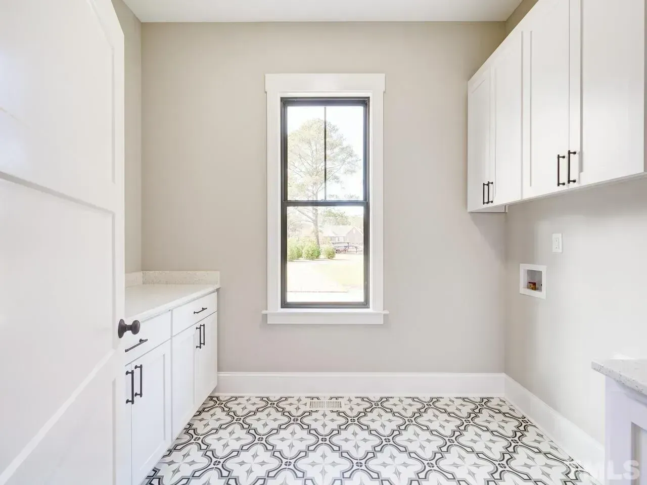 A bright, neutral-toned laundry room with white cabinets, a window, and patterned geometric floor tiles.